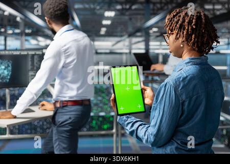 Black female specialist reviewing isolated copy space on tablet, updating data center infrastructure. Young engineer using mockup in server hub workspace filled with networking tech. Stock Photo