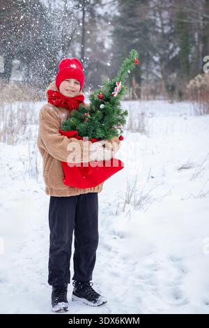 Smiling child holding small decorated Christmas tree in snowy winter forest with falling snow and festive holiday atmosphere. Stock Photo
