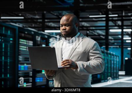 Technician in data center updating systems using laptop, protecting rigs from damaging software errors. African american man in server hub using notebook, looking for bugs causing gear to slow down Stock Photo