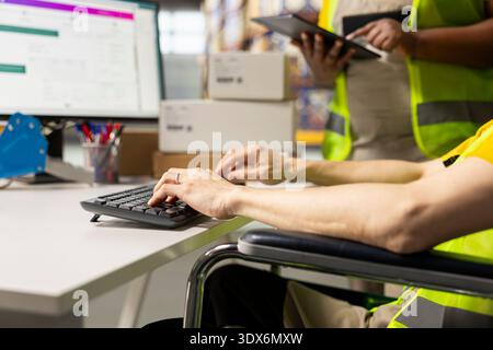 Industrial worker with disability uses airway bill numbers for tracking info on software, monitoring the order parcel status in a logistics hub with a colleague. Managing shipment operations. Stock Photo