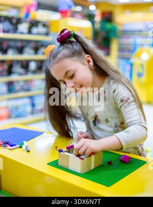 Girl concentrating while assembling colorful building bricks on green baseplate at yellow table in toy shop. Stock Photo