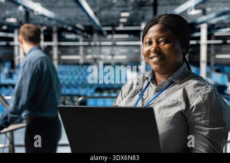 Close up of smiling worker in server farm next to colleague upgrading hardware for increased storage capacity. Jolly african american data center admin and coworker doing maintenance Stock Photo