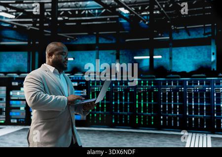 Data center software developer using laptop to protect servers from exploitable software vulnerabilities, auditing systems. African american server farm employee looking for bugs slowing down rigs Stock Photo