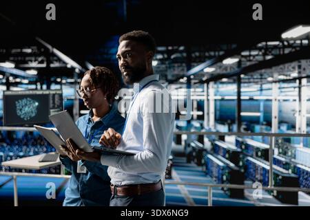 African american IT experts on industrial platform analyze server rigs algorithms in data center, using laptop and tablet. Server room IT specialists collaborating on finetuning PC configurations. Stock Photo