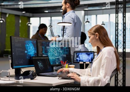 Female computer scientist in office relies on AI to run tasks across multiple devices, monitoring progress on screens. Young tech startup worker doing internship uses neural networks to develop AI Stock Photo