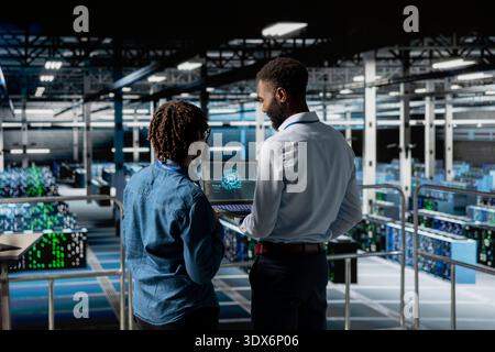 Server hub teamworking IT experts visualizing large language model architecture using laptop on elevated industrial platform. Data center system administrators collaborating on tasks. Stock Photo