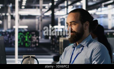 Smiling data center technician using PC to monitor neural network AI LLM visualization. Server room worker using computer to oversee rigs powering machine learning processes, camera A Stock Photo