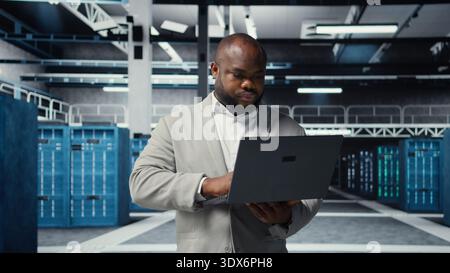 Technician walking in data center, using laptop to check hardware for system stability. Worker overseeing server farm, using notebook to configure infrastructure, optimizing performance, camera A Stock Photo