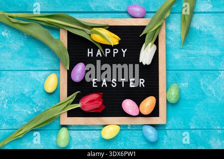 A festive flat lay Easter composition featuring a black letter board with white text reading Happy Easter surrounded by colorful glitter Easter eggs i Stock Photo