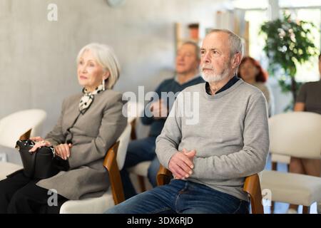 Focused elderly man wearing ordinary clothes sitting on chair with group of other students of different ages listening speech in light conference room Stock Photo