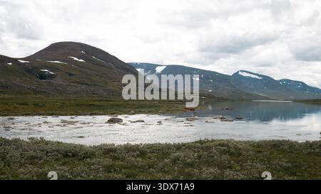 Sweden - July 7 2024: Mountain landscape with a calm lake, rocky shores, and distant peaks capped with snow under an overcast sky. A small red boat is Stock Photo