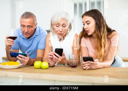 Elderly parents and adult daughter surf the internet using mobile phones in kitchen Stock Photo