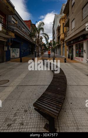 Two people are walking down a street with a curved wooden bench in the foreground. Stock Photo