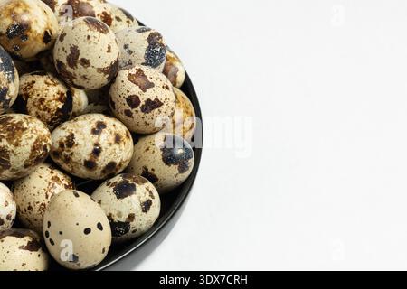 Many quail eggs are stored in a dark bowl placed on a white surface. These eggs have various patterns and colors. T Stock Photo
