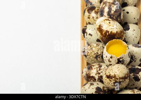 A collection of quail eggs is placed on a wooden tray. One egg is broken, showing its yellow yolk. The eggs have dark spots on a light shell. Stock Photo