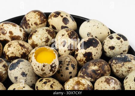 Many quail eggs are gathered on a black plate. One egg is cracked open, revealing a yellow yolk inside. Stock Photo