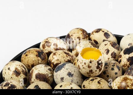 Small quail eggs are arranged in a bowl. Most eggs remain whole, while one egg has a broken shell and reveals a bright yellow yolk inside. Stock Photo