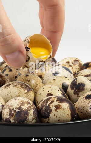 A hand reaches for a quail egg that is cracked open. The yolk is visible inside the shell. Other quail eggs lie in a bowl. Stock Photo