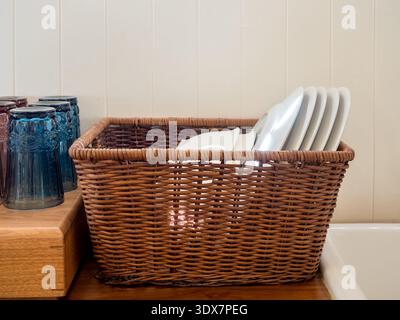Clean white bowls and dishes neatly arranged in vintage wicker basket on wooden kitchen counter. Concept of home organization, dish storage, and kitch Stock Photo