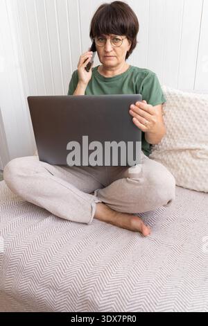 Mature woman working remotely on laptop while talking on phone Stock Photo