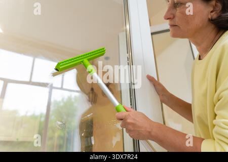 Mature female cleaning windows with green squeegee at home. Cleaning and professional house cleaning, small business Stock Photo
