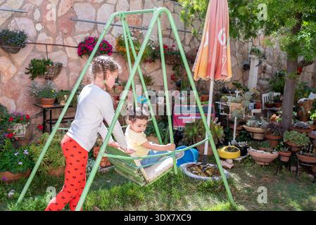 Two sisters are playing on a swing in their backyard filled with colorful plants and flowers. They share smiles and laughter as they enjoy the warm sunshine together. Stock Photo