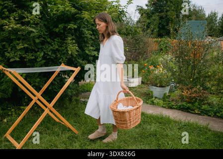 Young woman carrying wicker laundry basket walking towards wooden clothesline in a lush green garden, preparing to hang clean laundry to dry on a sunny day Stock Photo