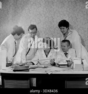Vintage black and white archival photograph capturing a dedicated team of five medical professionals in classic white lab coats gathered around a desk to carefully examine an electrocardiogram printout. The doctors and nurses are having a serious clinical consultation inside an office at City Hospital No 2 in Sloviansk, Ukrainian SSR during the 1980s. They study a long EKG paper strip over a table covered with documents, against typical Soviet floral wallpaper, highlighting everyday healthcare teamwork in peaceful Donbas. Stock Photo