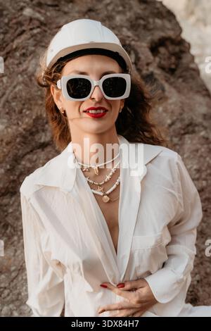 Woman hard hat sunglasses posing in a white shirt and safety helmet against a rocky backdrop for a creative concept Stock Photo