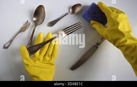 Close-up of hands in yellow rubber gloves scrubbing cutlery, spoons and forks, with purple sponge on a wet white surface. Concept of hygiene, disinfection, hand protection, and routine house cleaning. Stock Photo