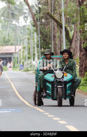 Authentic Thailand: Thai gardening workers ride along a rural road in southern Thailand in Mai Khao district on a green electric cargo Rutrike Voyage Stock Photo
