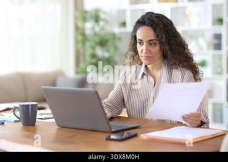 Serious freelance worker holding report and checking laptop working at home Stock Photo