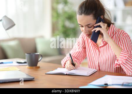 Serious self employed talking on phone writing in agenda working at home Stock Photo