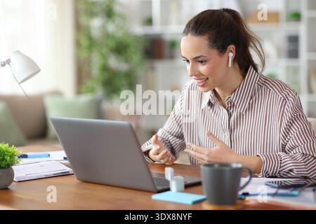 Happy tele worker making video call using earbuds and laptop working in a house interior Stock Photo