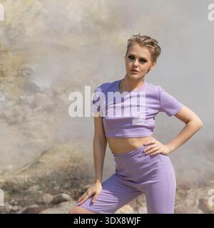 A stylish young woman with a pixie haircut and bold makeup wears a lavender two-piece set. She poses confidently outdoors against a blurred, steamy ge Stock Photo