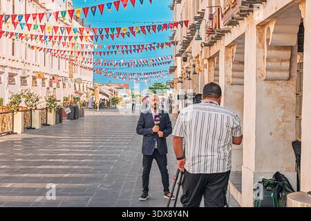 1 December 2025, Doha, Qatar: Reporter broadcasting live on television, holding a microphone while team films during event celebration in Souq Waqif Stock Photo