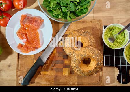 Two seeded bagels on wooden cutting board beside ramekin of mashed avocado, salmon and spinach salad in background. Concept of healthy breakfast, meal prep and fresh ingredients Stock Photo