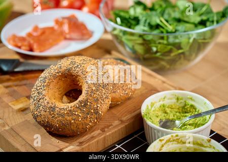Two seeded bagels on wooden cutting board beside ramekin of mashed avocado, salmon and spinach salad in background. Concept of healthy breakfast, meal prep and fresh ingredients Stock Photo