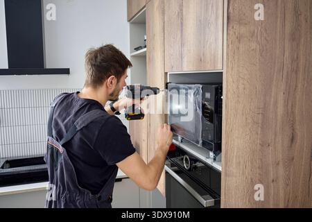 Man using cordless screwdriver to fixing new built in microwave oven with screws in modern kitchen cabinet. Concept of home appliances installing Stock Photo
