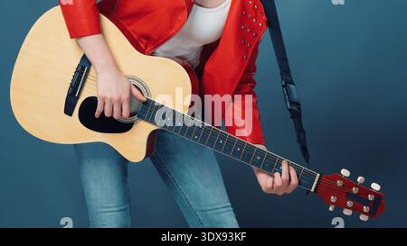 Musician in a red studded jacket and jeans plays an acoustic guitar. Cropped view with vibrant colors on a blue background. Concept for music lessons, Stock Photo