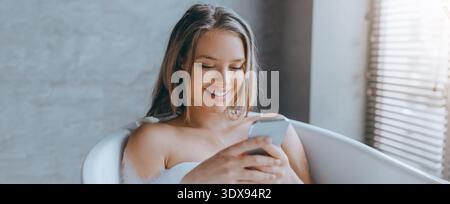 Woman Using Smartphone While Relaxing in Bathtub With Natural Light in Background Stock Photo
