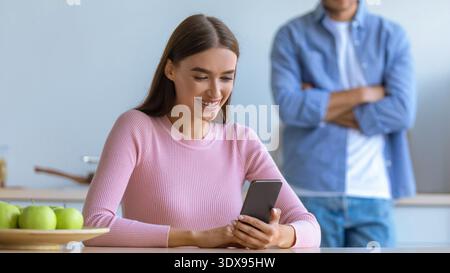 Woman Smiles While Using Smartphone in Kitchen With Man Standing Behind Her Stock Photo
