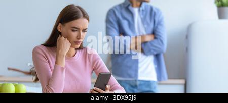 Young Woman Using Smartphone While Man Stands Nearby With Crossed Arms at Home Stock Photo