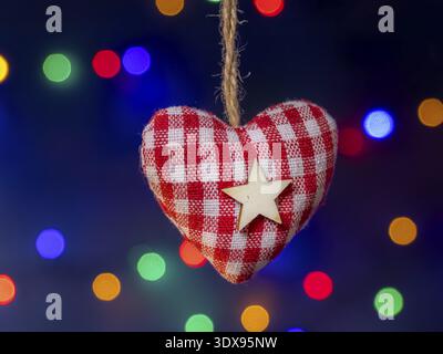 A fabric heart ornament in red and white gingham with a wooden star, hanging against festive multicolored lights. Warm, cozy holiday decor that convey Stock Photo