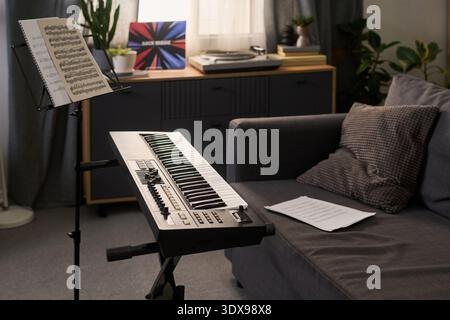 Electronic keyboard standing in living room near sofa with sheet music on stand and paper on floor, vinyl record player and decorative plants in background, no people visible Stock Photo