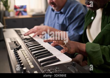 Middle aged Caucasian man and middle aged Black man playing electronic keyboard together, focusing on hands pressing keys, collaborating on music in indoor setting Stock Photo