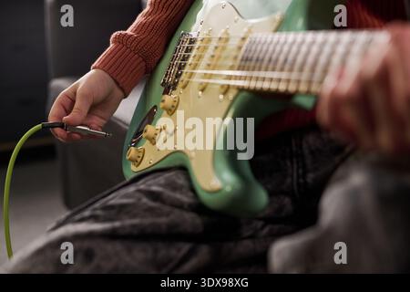 Caucasian teenager plugging electric guitar cable into instrument, sitting indoors with focus on hands and guitar body, demonstrating preparation for playing music Stock Photo
