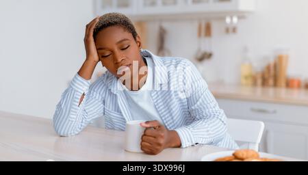 Woman Sitting at Kitchen Table Holding Mug, Looking Tired and Thoughtful, Morning Time Stock Photo