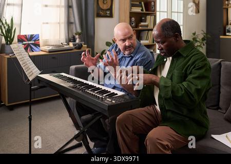 Middle aged Caucasian man and senior Black man sitting on sofa playing electronic keyboard together, gesturing with hands while reading sheet music, engaging in collaborative music practice Stock Photo