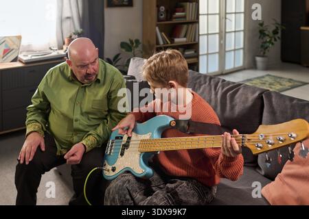 Caucasian middle aged man teaching Caucasian boy playing electric bass guitar on sofa in living room, man gesturing while boy focusing on instrument, natural indoor setting Stock Photo
